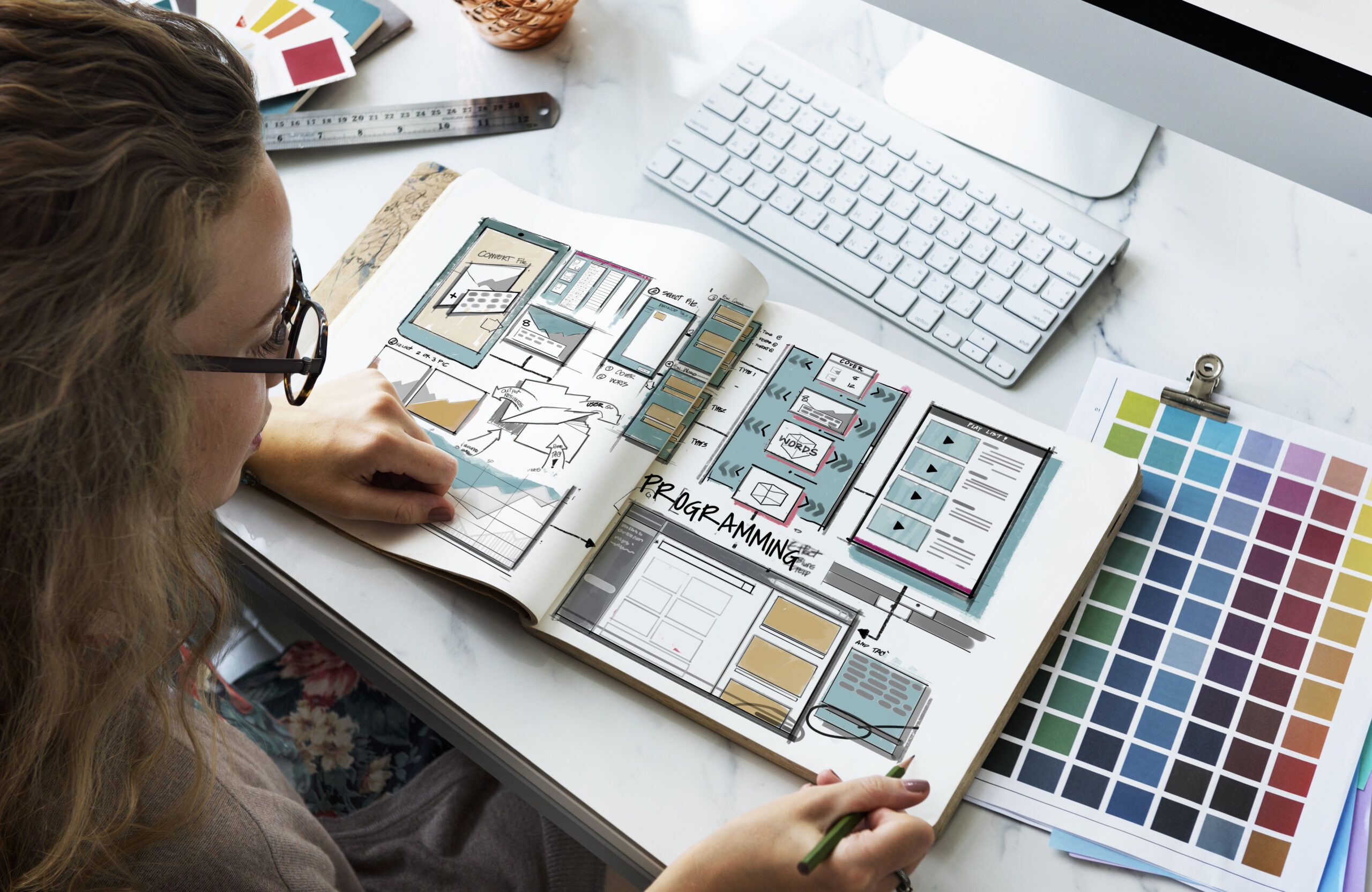 Mulher trabalhando em uma mesa com cadernos de esboços de design, cercada por paleta de cores, computador e materiais de desenho.
