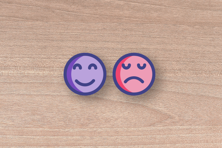 Dois ícones emoji sobre madeira, um sorrindo e roxo, outro triste e vermelho, representando emoções opostas.