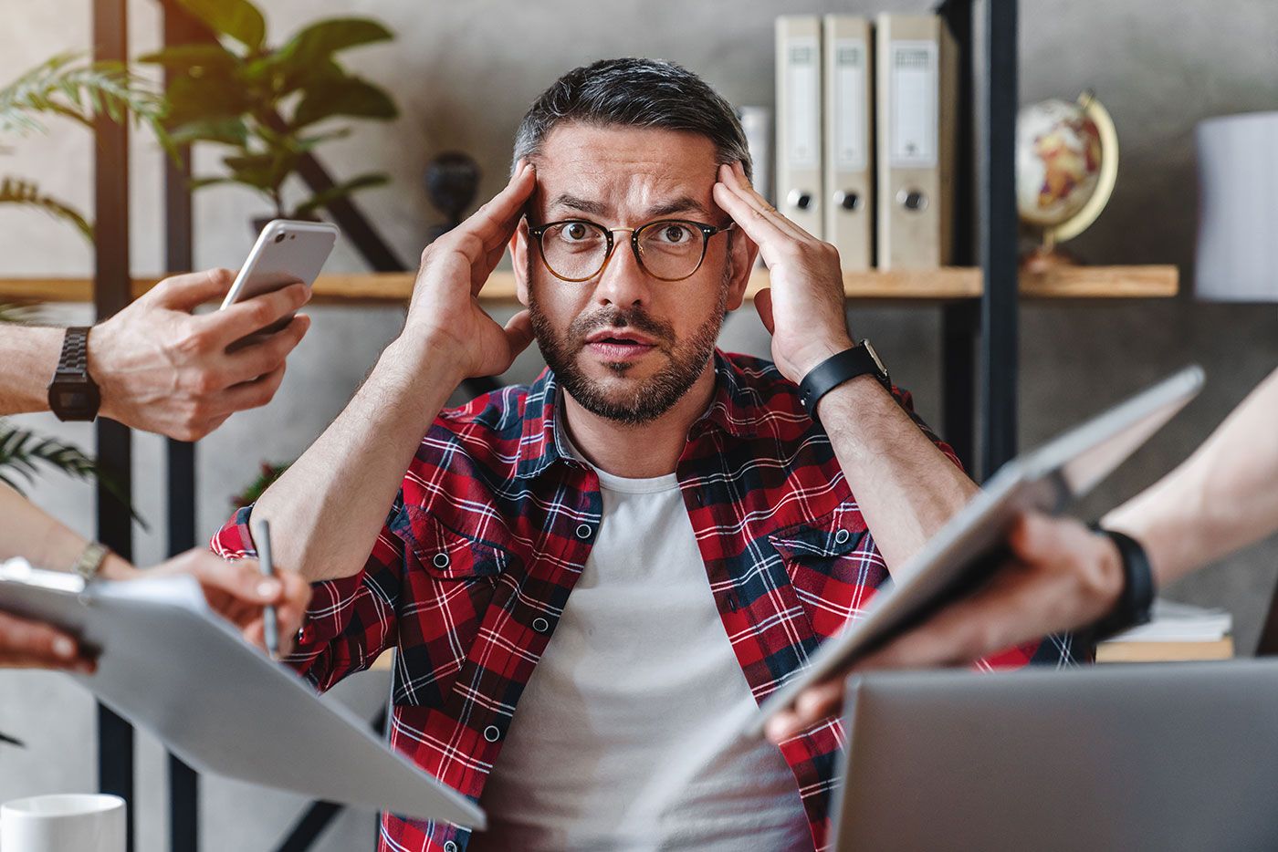 Homem estressado em escritório, segurando a cabeça com as mãos, cercado por colegas com dispositivos e papéis.