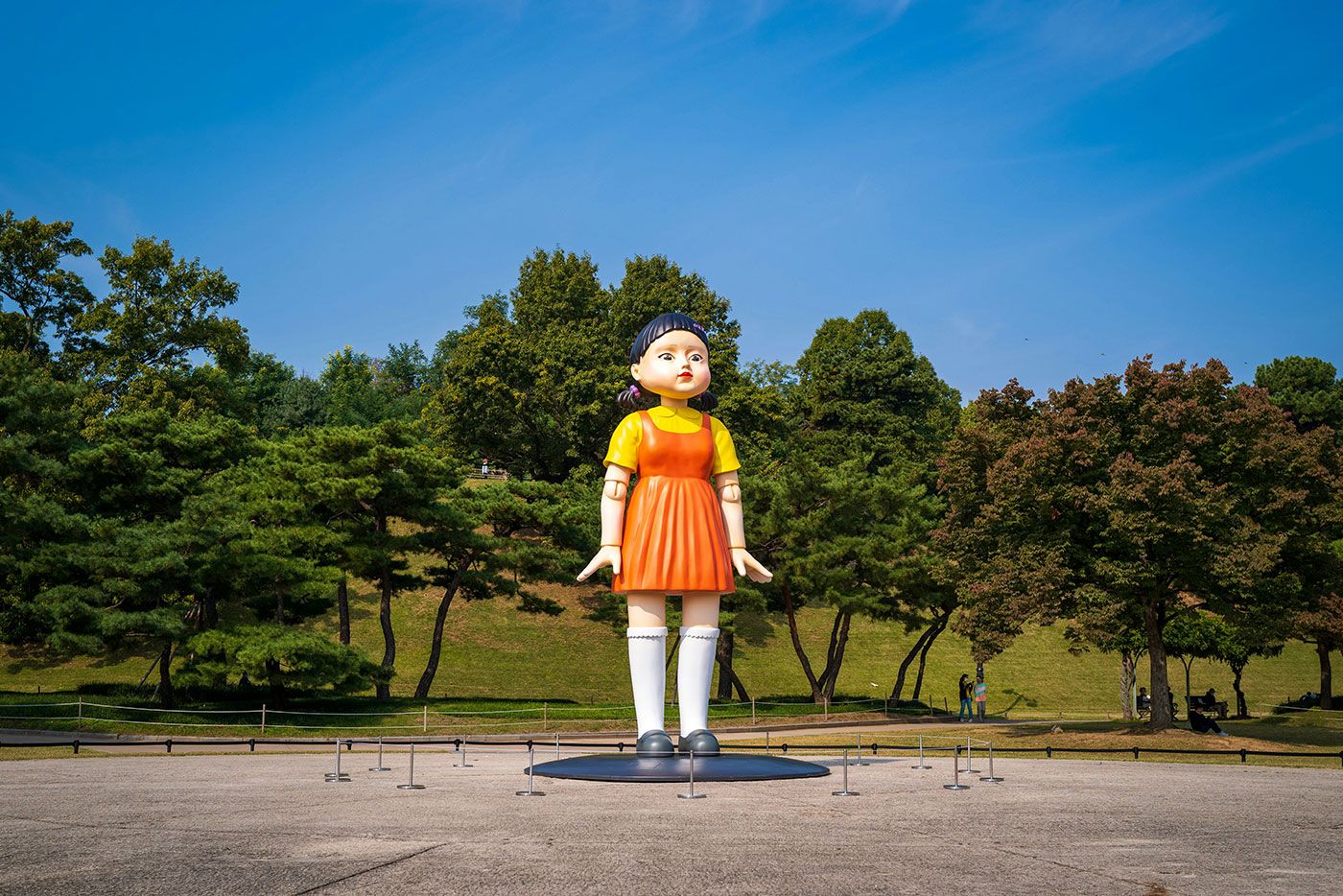 Escultura gigante de uma menina em vestido laranja posa em um parque com céu azul e árvores ao fundo.
