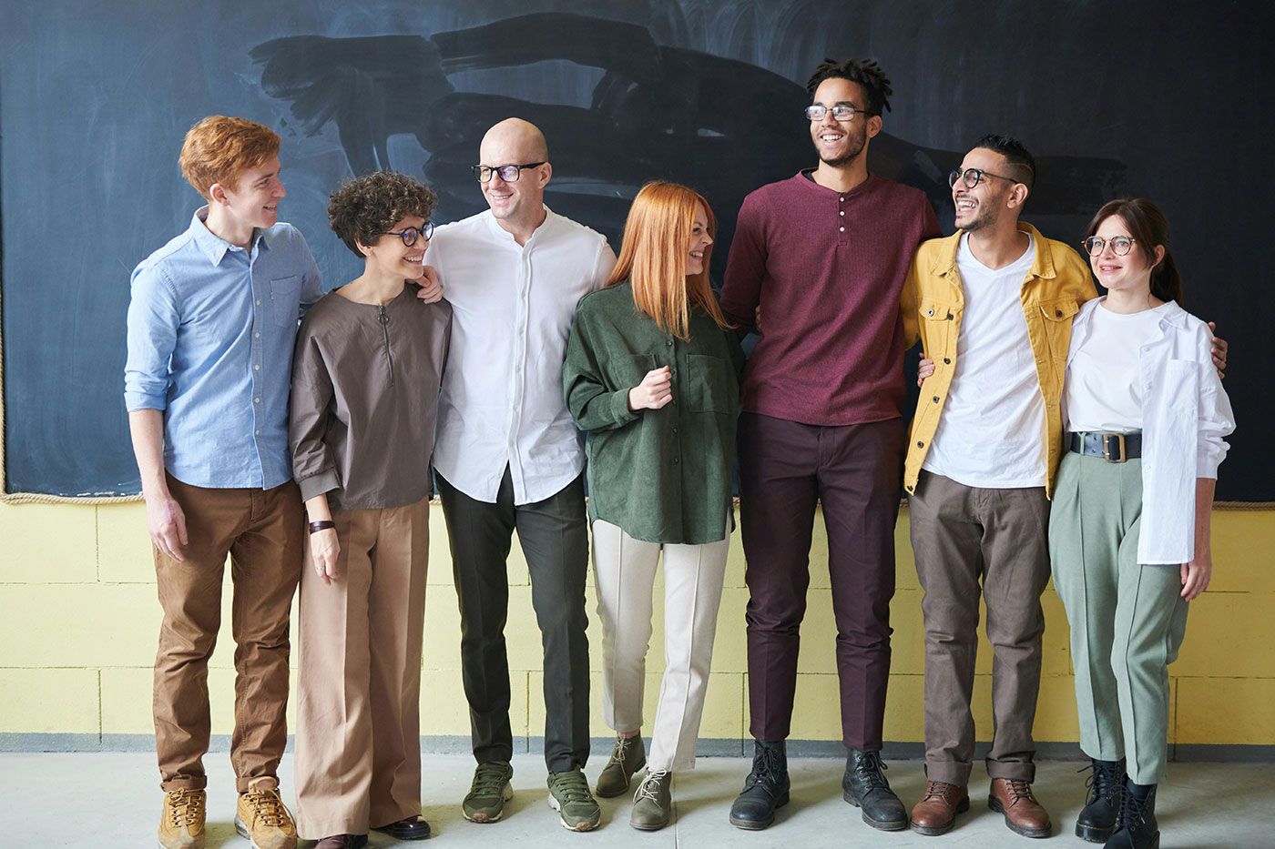 Grupo diversificado de sete pessoas sorrindo e se abraçando em frente a uma lousa escolar. Eles parecem alegres e descontraídos.
