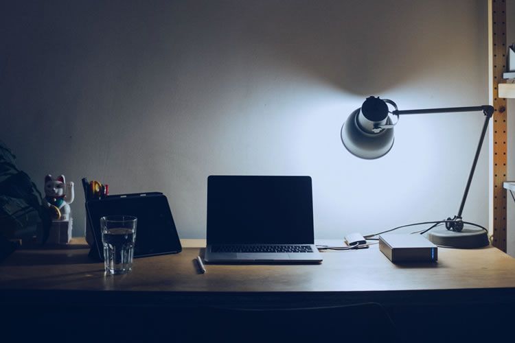 Mesa de trabalho iluminada por uma luminária, com um laptop, tablet, copo d'água e pequenos objetos decorativos. Ambiente sombrio com paredes claras.