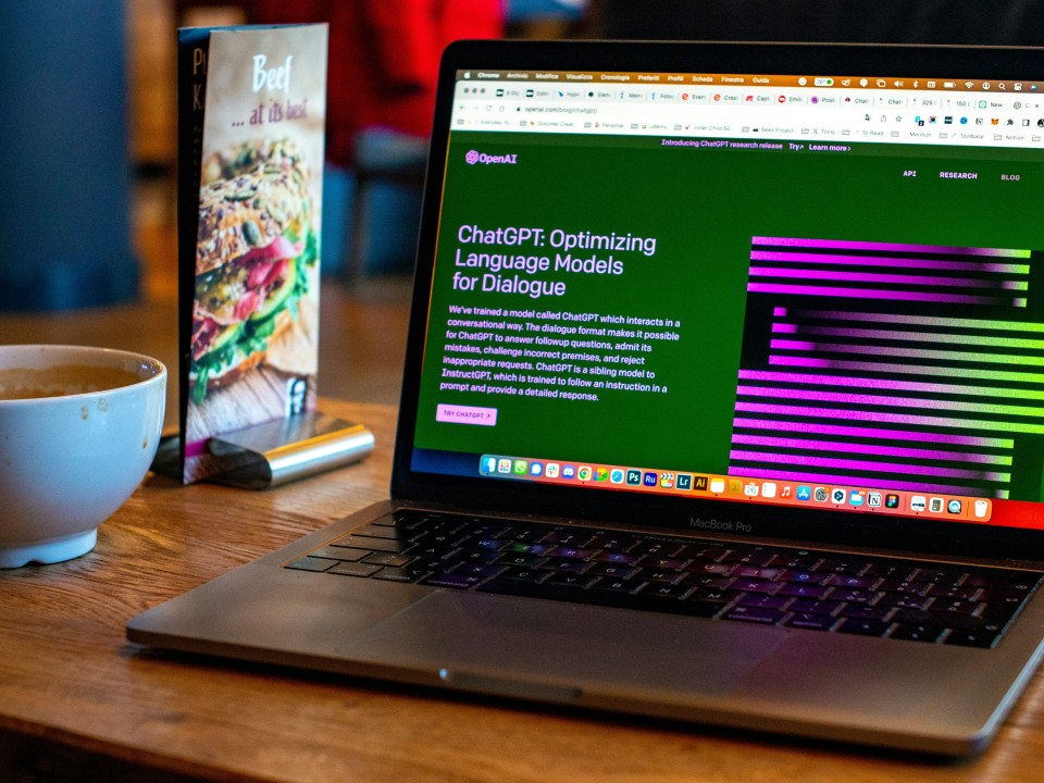 Laptop aberto mostrando site da OpenAI sobre ChatGPT em mesa de café, ao lado de uma xícara e um celular sob um folheto de menu.