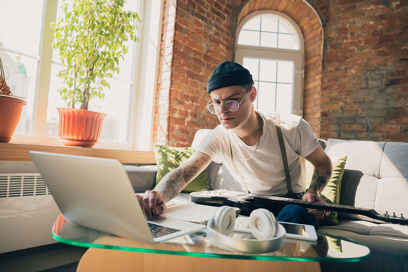 Homem jovem com tatuagens usando laptop e segurando guitarra sentado em sofá em ambiente iluminado com tijolos à vista.