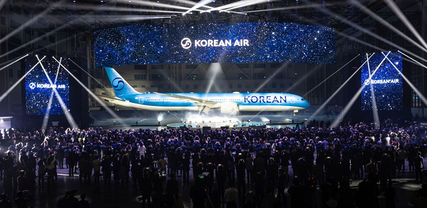Evento da Korean Air em hangar iluminado, com multidão, avião azul e painéis de LED exibindo o logo da companhia.