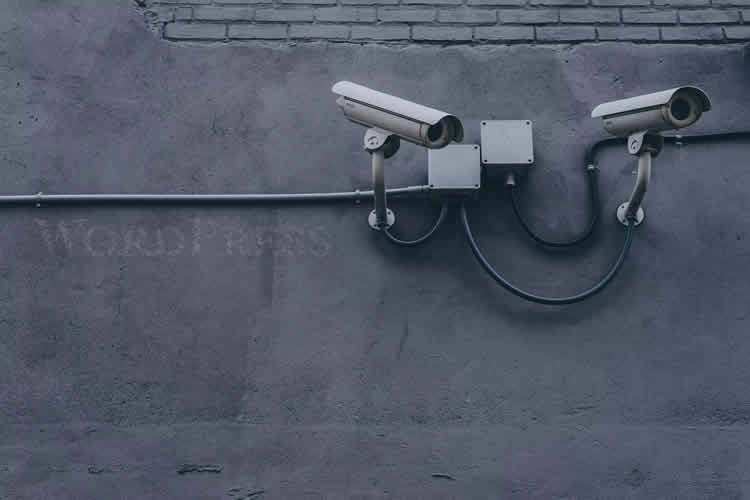 Duas câmeras de vigilância estão montadas em um muro de tijolos cinza, com a palavra 