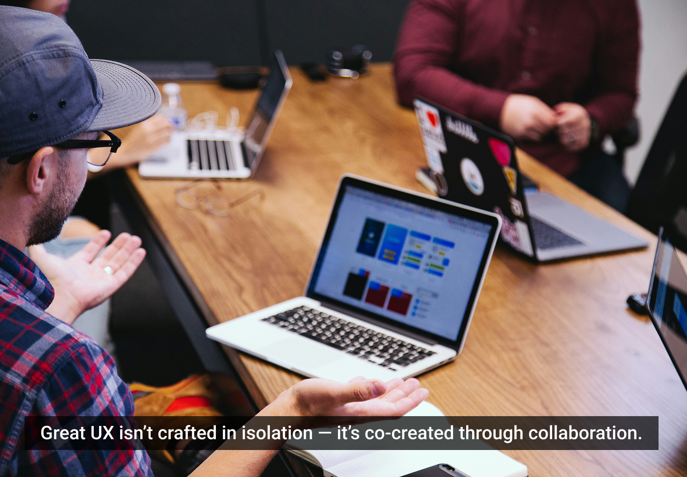 Três pessoas colaboram em torno de laptops em uma mesa de escritório, discutindo um projeto de design de interface do usuário visível na tela de um computador.