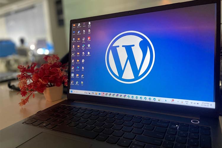 Laptop com tela exibindo o logo do WordPress em um ambiente de escritório com flores vermelhas desfocadas ao lado.