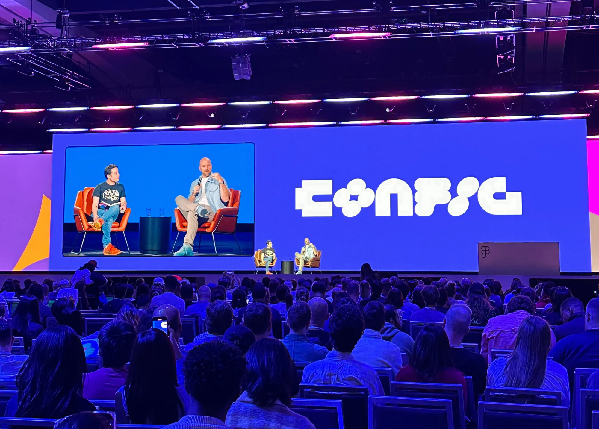 Duas pessoas sentadas em cadeiras laranjas em um palco de conferência, dialogando diante de uma grande audiência, com um grande painel digital ao fundo exibindo o logo 