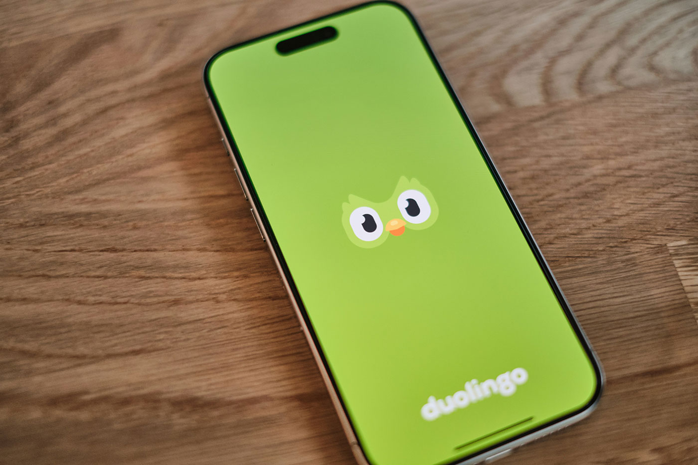 Smartphone sobre uma mesa de madeira exibindo a tela inicial do aplicativo Duolingo, com fundo verde e um desenho de coruja.