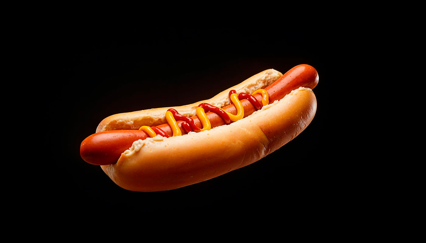 Close-up de um cachorro-quente com salsicha e condimentos sobre fundo preto.