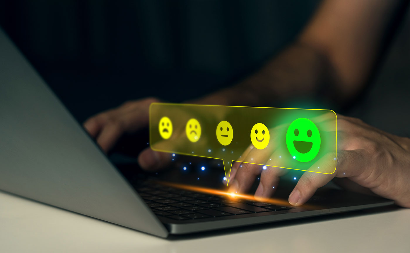 Mãos sobre um teclado de laptop mostrando uma balão de avaliação holográfico com emojis saindo do triste até o muito feliz.
