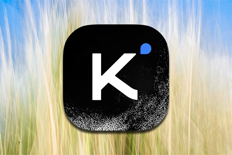 Ícone preto com a letra 'K' branca e um ponto azul, sobre fundo desfocado que simula textura de grama ou campo.