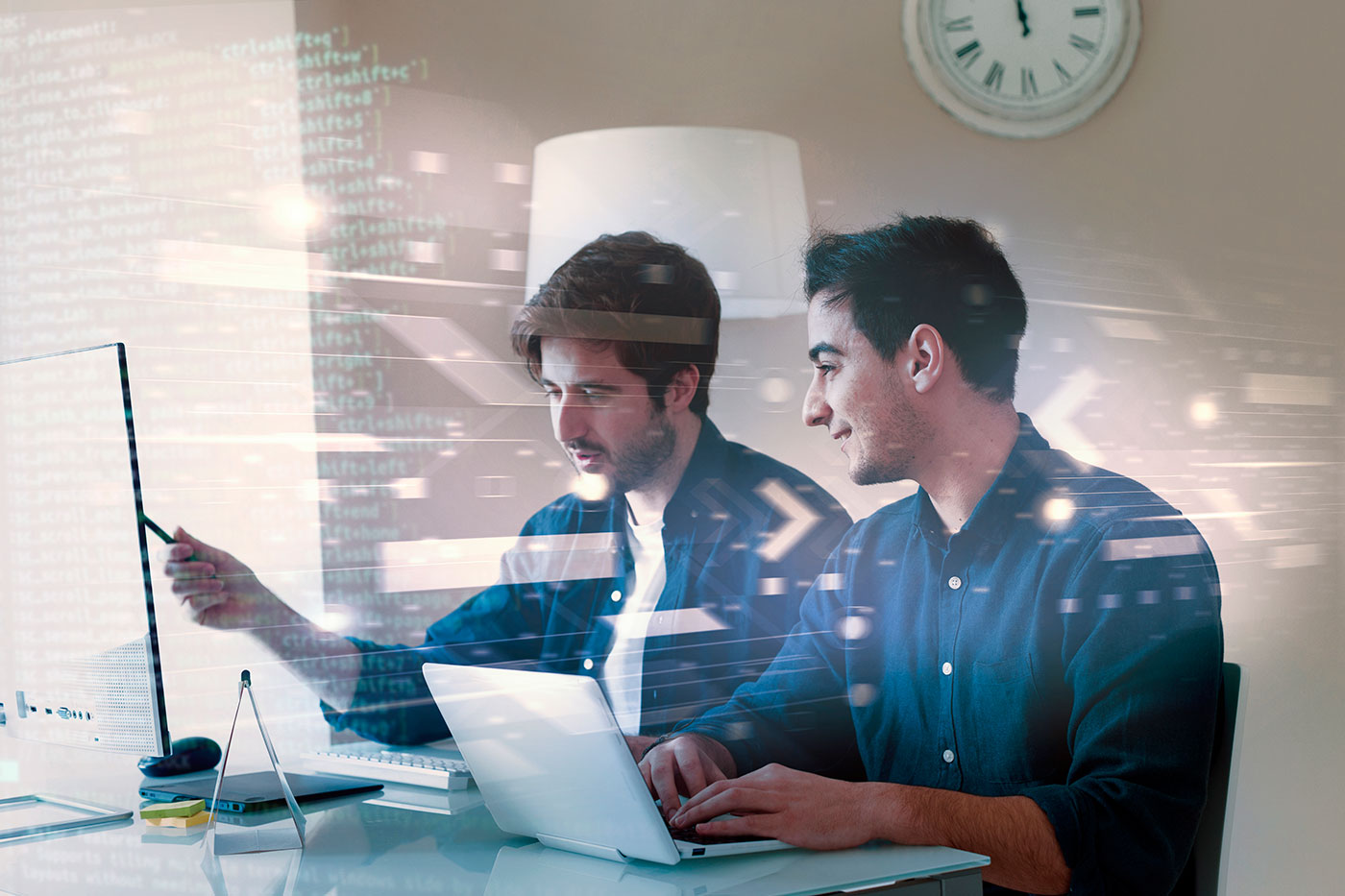 Dois homens jovens trabalham lado a lado em computadores em um ambiente de escritório, com códigos de programação visíveis ao fundo.