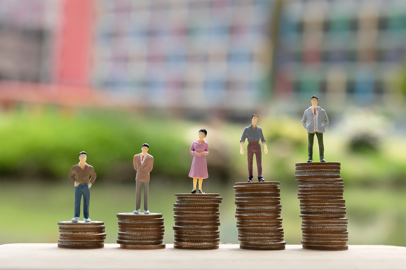 Miniaturas de pessoas em diferentes trajes de pé em pilhas crescentes de moedas, simbolizando níveis variados de renda ou sucesso financeiro, ao fundo, edifícios desfocados.