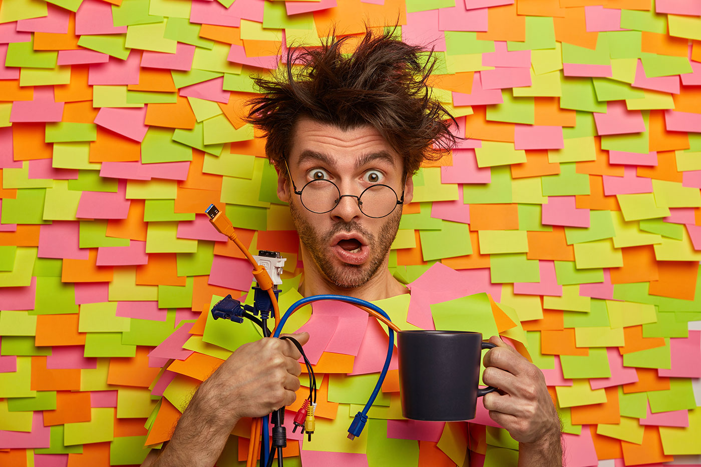 Homem surpreso com cabelo despenteado e óculos redondos segura uma caneca e cabos, cercado por coloridos papéis adesivos em fundo laranja e verde.